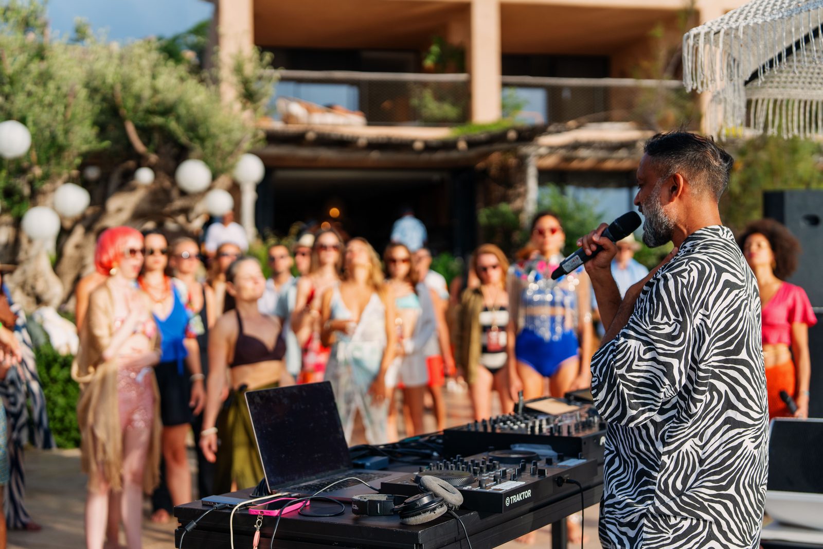 Niraj Naik speaking into a microphone behind DJ decks while addressing a crowd of participants at an outdoor breathwork event