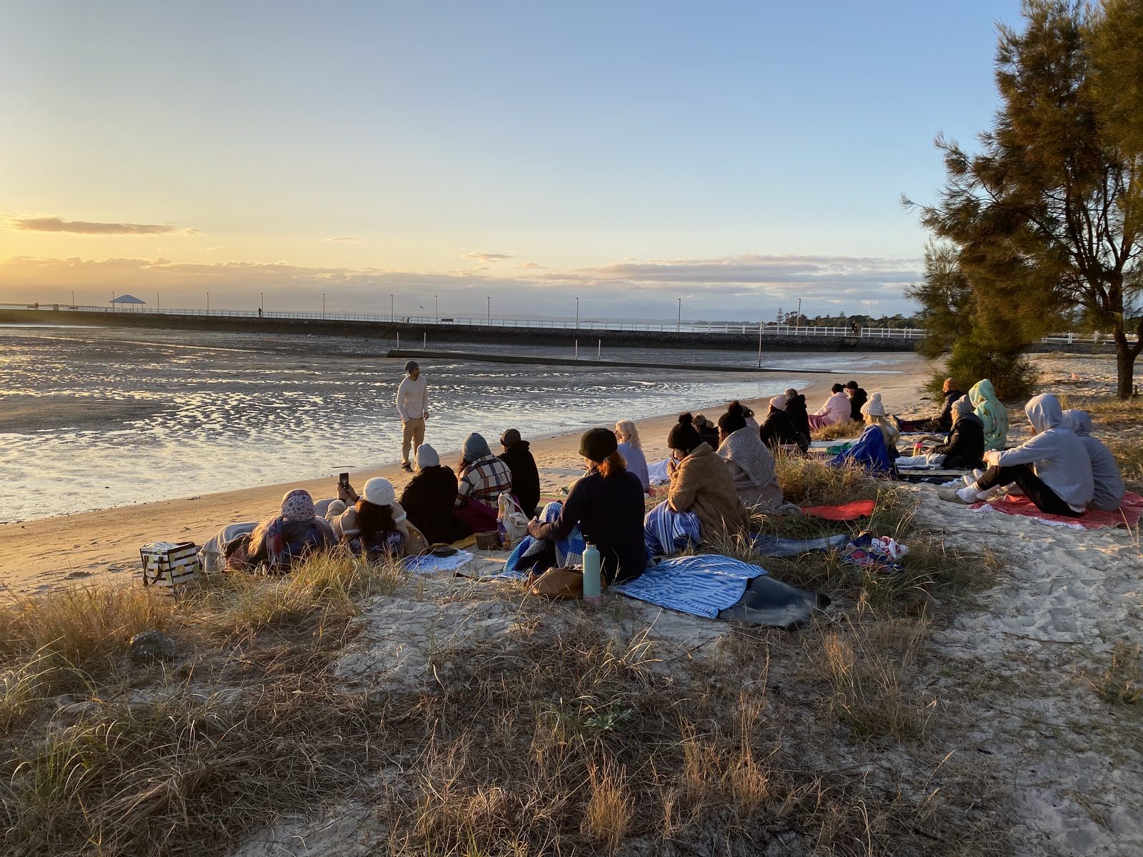 Group wrapped in blankets sitting on a beach at sunrise while a facilitator guides them through a breathing practice