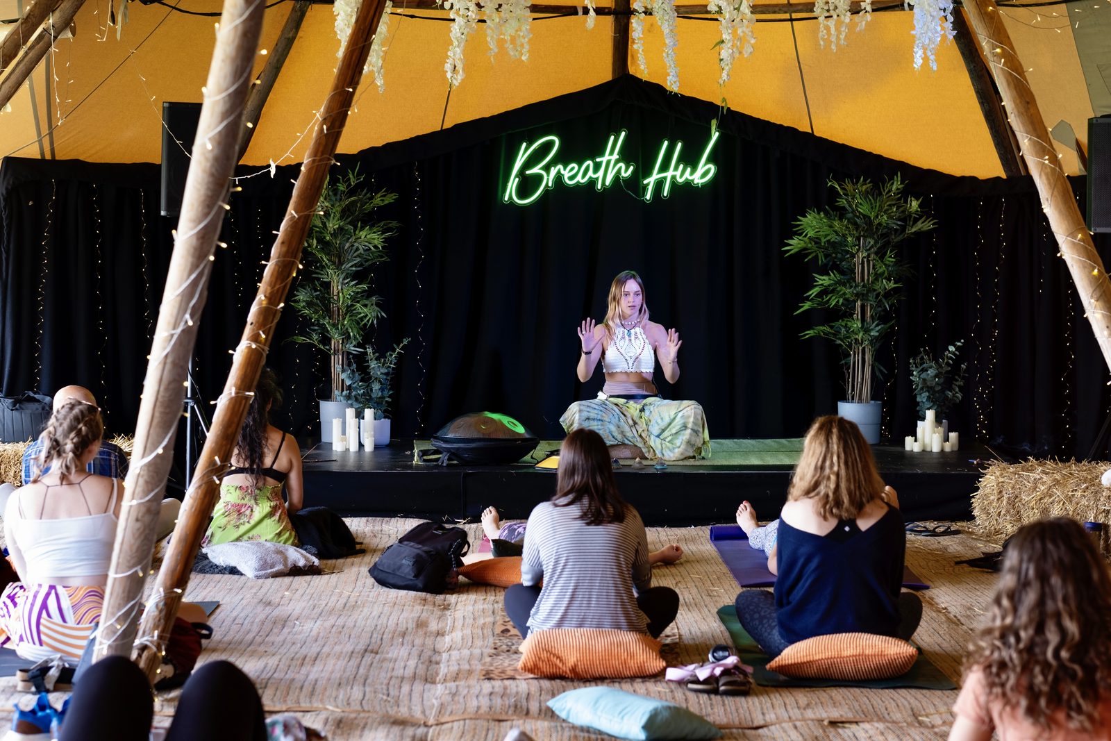 Breathwork instructor sitting cross-legged with both hands raised, demonstrating a breathing pattern to a small group seated on cushions under a tipi