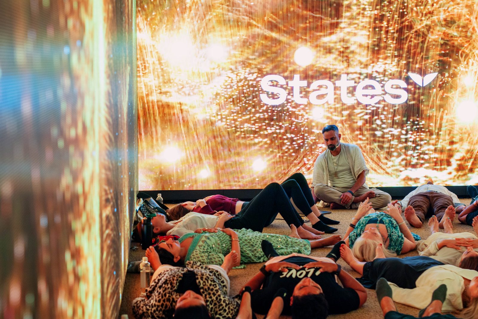 Niraj Naik sitting cross-legged at the front of a large guided breathwork session while participants lie on the floor during the integration phase