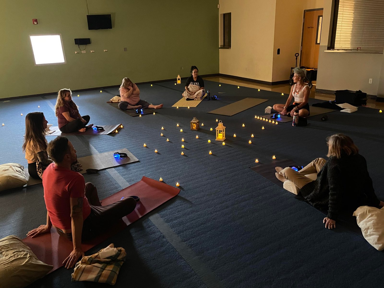 Nine people sitting on mats in a candlelit room during a SOMA Breath class, wireless headphones on the floor beside each person